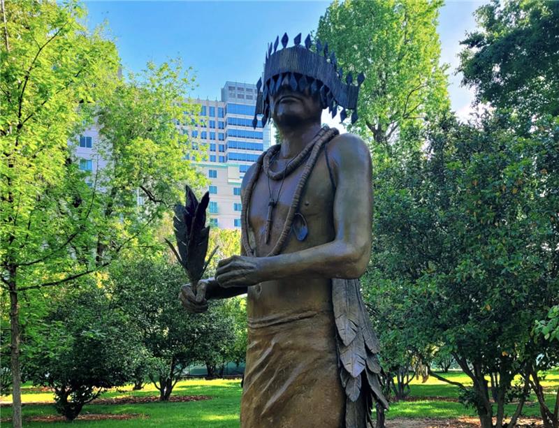 Bronze statue of a Native American in a wooded park.